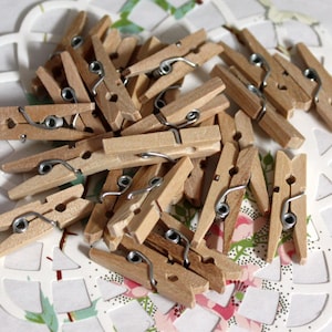 May include: A collection of small, natural wood clothespins with silver-colored metal springs. The clothespins are scattered on a white, decorative paper doily with a floral pattern. The wood has a light brown color.