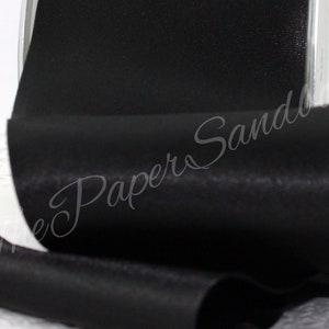 May include: A roll of black satin ribbon. The ribbon is tightly wound and has a smooth, shiny surface. The image is a close-up, highlighting the texture and sheen of the fabric. The ribbon is likely used for crafts, gift wrapping, or decorative purposes.