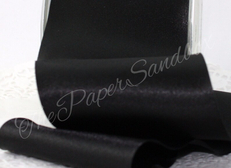 May include: A roll of black satin ribbon. The ribbon is tightly wound and has a smooth, shiny surface. The image is a close-up, highlighting the texture and sheen of the fabric. The ribbon is likely used for crafts, gift wrapping, or decorative purposes.