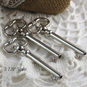May include: Three silver-colored decorative keys with ornate tops and keyhole ends are displayed. The keys are approximately 2 7/8 inches long. The keys are arranged on a white, lacy surface.