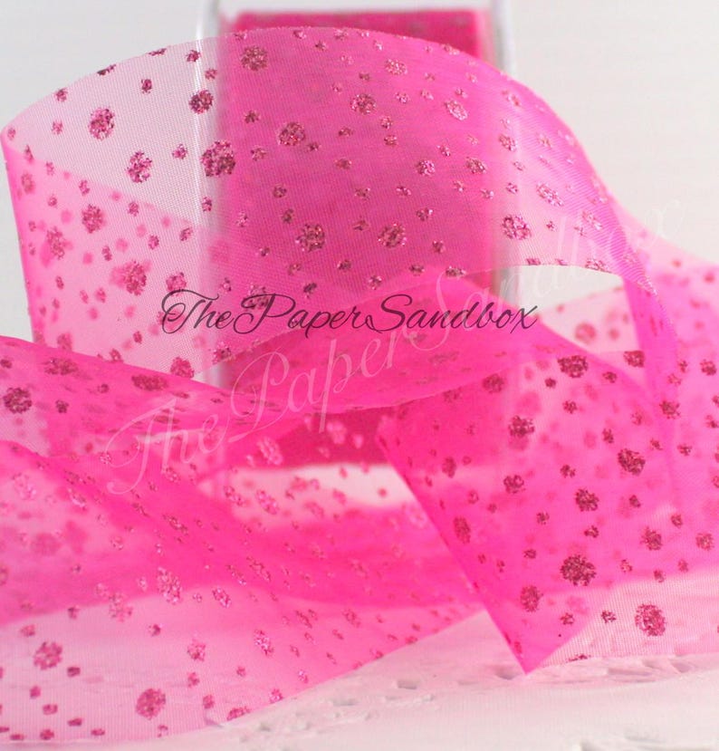 May include: Bright pink sheer ribbon with glitter accents. The ribbon has a dotted pattern of glitter and the text "The Paper Sandbox" is visible. This ribbon is suitable for crafts, gift wrapping, and decorations.