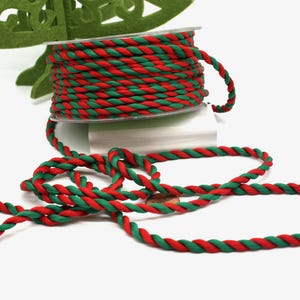 May include: A spool of red and green twisted rope sits on a white surface. The rope is unwound, with a length of it lying in loops. A green felt Christmas tree is in the background.
