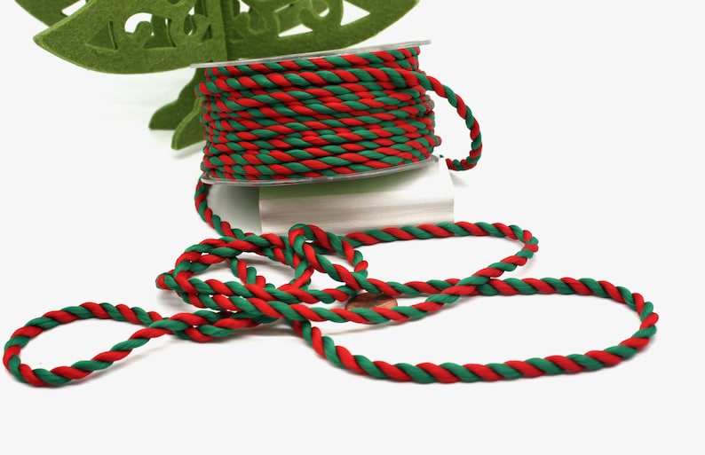 May include: A spool of red and green twisted rope sits on a white surface. The rope is unwound, with a length of it lying in loops. A green felt Christmas tree is in the background.