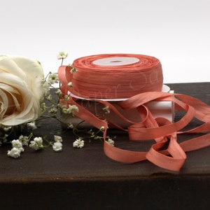 May include: A roll of coral-colored ribbon sits on a white spool, with loose ribbon trailing. A white rose and sprigs of white baby's breath are arranged nearby. The ribbon is likely made of silk or a similar material, perfect for crafts and gift wrapping.