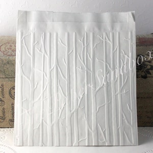 May include: A white paper gift bag with an embossed forest design. The design features vertical tree trunks and branches. The bag is rectangular with a folded top edge. The words "Paper Sandbox" are visible in the design.