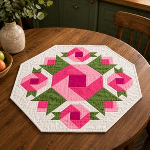 May include: An octagonal quilted table topper featuring a geometric pattern in shades of hot pink, light pink, and olive green on a white background. The design consists of a central square surrounded by four stylized floral motifs.