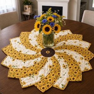 May include: A circular quilted table topper featuring a spiral pattern of yellow and white fabric with sunflower prints. The center has a dark brown circular patch with a small sunflower motif. A glass vase of sunflowers sits in the middle.