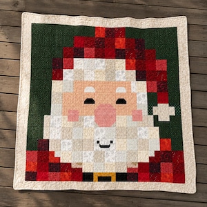 May include: A square quilt featuring a pixelated Santa Claus face design. The quilt displays a red hat, a white beard, a pink nose, and a dark green background. It is framed by a cream-colored border and finished with visible grid-like stitching.