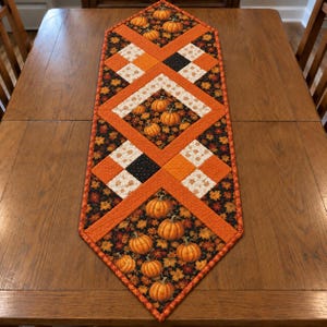 May include: A quilted table runner featuring a patchwork design with orange, black, and white fabric. The pattern includes sections of black fabric with orange pumpkin prints, solid orange borders, and white squares with small orange leaf motifs.