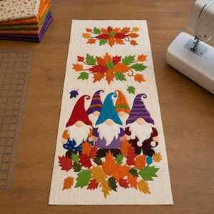 May include: A rectangular fabric table runner featuring a cream-colored background with appliqued designs of three gnomes wearing colorful hats and white beards, surrounded by vibrant autumn leaves in shades of red, orange, yellow, and green.
