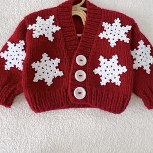 May include: A red knitted cardigan with white snowflake appliqués. The sweater has a V-neck, three white buttons, and long sleeves. The garment is displayed on a wooden hanger.