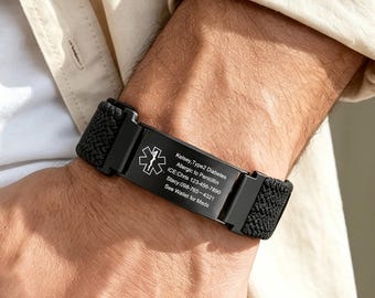 Puede incluir: Una pulsera de alerta médica negra con una banda de tela tejida y una placa metálica rectangular. La placa muestra el símbolo de la Estrella de la Vida y texto grabado que incluye información médica, contactos de emergencia y la frase See Wallet for MDI.