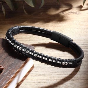 May include: Black leather bracelet with silver-colored beads and accents. The bracelet features a magnetic clasp and is displayed on a wooden surface. A stylish accessory for everyday wear.