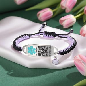 Könnte beinhalten: Ein schwarz-lilafarbenes medizinisches Armband mit einer silbernen Platte. Die Platte hat ein medizinisches Symbol, einen QR-Code und die Worte "HEALTH INFO" und "SCAN ME". Ein kleiner, runder, lila Anhänger hängt am Armband.