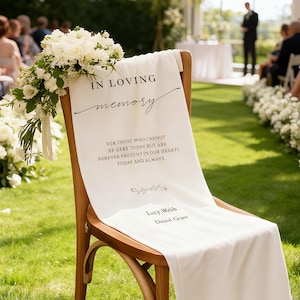 Puede incluir: Un camino de silla de tela blanca con la inscripción "IN LOVING memory" en escritura negra. Incluye un mensaje conmemorativo y nombres. Un ramo de flores blancas adorna la silla, en un entorno de jardín.
