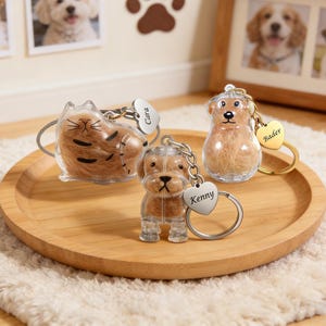 Puede incluir: Tres llaveros con temática de mascotas en una bandeja de madera. Uno es un gato y dos son perros. Cada llavero tiene un exterior de plástico transparente con un interior esponjoso. Cada llavero tiene una etiqueta en forma de corazón con un nombre.