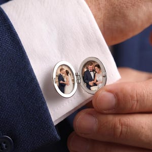 Op de afbeelding: Een zilverkleurige, ovale medaillon-manchetknop bevestigd aan een wit overhemdmanchet. Het medaillon staat open en toont twee kleine, ronde foto's: één met een omhelzend stel en de andere met een stel tijdens een huwelijksceremonie.