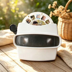 Puede incluir: Un visor blanco de estilo retro con un ocular negro y un disco giratorio que muestra fotos familiares. El visor está sobre una superficie de madera, con una cesta y vegetación en el fondo. Un artículo nostálgico para ver recuerdos.