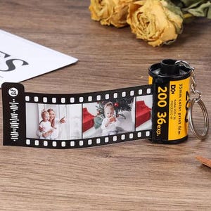 Pode incluir: Um porta-chaves de tira de filme preta com três ranhuras para fotos e um cartucho de filme colorido de 35 mm. A tira de filme exibe fotos de família. O cartucho é amarelo e preto com o texto "200 36exp."