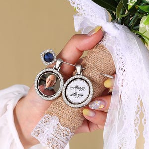 Op de afbeelding: Een bruidsboeket met witte rozen en groen, vastgehouden met een met jute omwikkelde handgreep versierd met kant en twee herdenkingsbedels. Eén bedel heeft een foto, de andere luidt "Always with you."