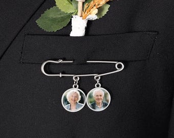 Puede incluir: Un broche de imperdible plateado sujeto a una chaqueta de traje negra. El broche cuenta con dos dijes circulares que muestran el retrato de una persona cada uno, y está adornado con un boutonniere floral blanco con hojas verdes.