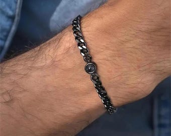 Puede incluir: Una pulsera de cadena negra con un acento circular central. La pulsera está hecha de eslabones entrelazados y se usa en una muñeca. El acento parece ser una piedra de color oscuro.