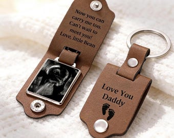 May include: Two brown leather keychains with silver metal rings and snap buttons. One keychain is open, revealing a black and white ultrasound image and text reading: Now you can carry me too. Can't wait to meet you! Love, little Bean. The other reads: Love You Daddy.