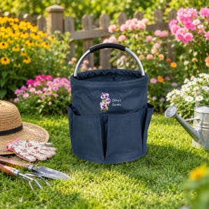 Puede incluir: Una bolsa de jard&iacute;n de lona azul marino con un asa de metal plateado y m&uacute;ltiples bolsillos exteriores. La parte frontal presenta una ilustraci&oacute;n de pensamientos morados y el texto blanco "Selina's Garden" sobre la tela.