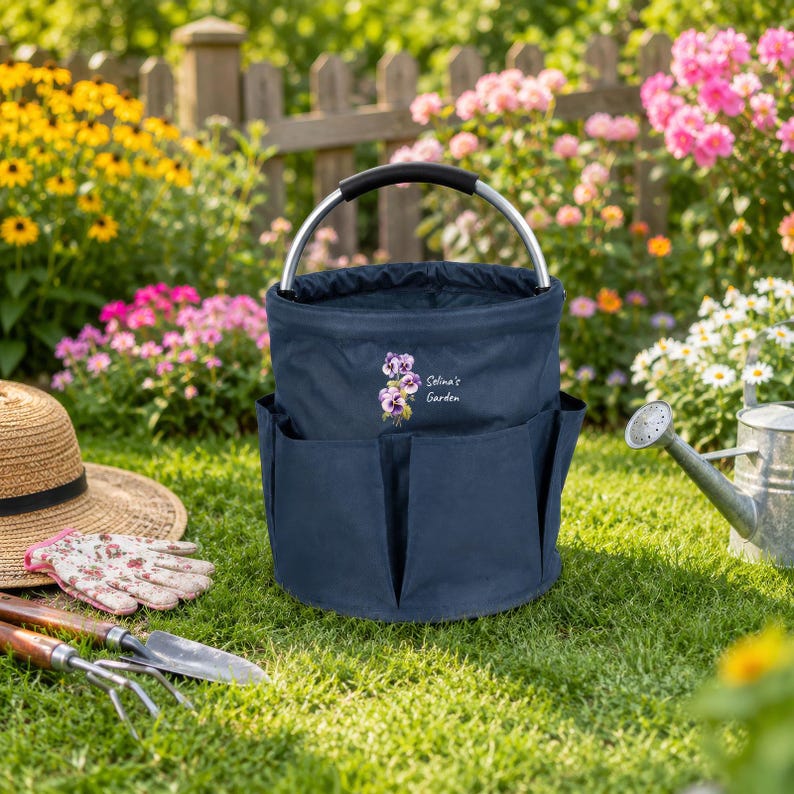 Puede incluir: Una bolsa de jard&iacute;n de lona azul marino con un asa de metal plateado y m&uacute;ltiples bolsillos exteriores. La parte frontal presenta una ilustraci&oacute;n de pensamientos morados y el texto blanco "Selina's Garden" sobre la tela.