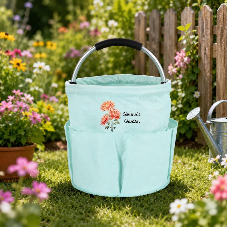 Puede incluir: Una bolsa de jard&iacute;n de tela en color verde menta claro con un asa de metal plateado y agarre negro. El frente presenta una ilustraci&oacute;n impresa de crisantemos naranjas y el texto negro Selina's Garden. Tiene varios bolsillos.