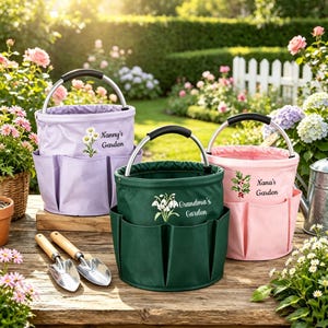 May include: Three cylindrical garden tool bags in lavender, dark green, and pink, each with a silver handle and exterior pockets. The lavender bag features a daisy graphic and text Nanny's Garden, the green bag has snowdrops and Grandma's Garden, and the pink bag has holly and Nana's Garden.