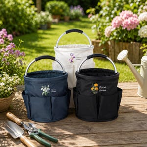 Puede incluir: Tres bolsas de jard&iacute;n cil&iacute;ndricas con asas de metal plateado y bolsillos exteriores. La bolsa azul marino muestra flores blancas y el texto "Mom's Garden", la blanca flores moradas y "Grandma's Garden", y la negra flores naranjas y "Selina's Garden".