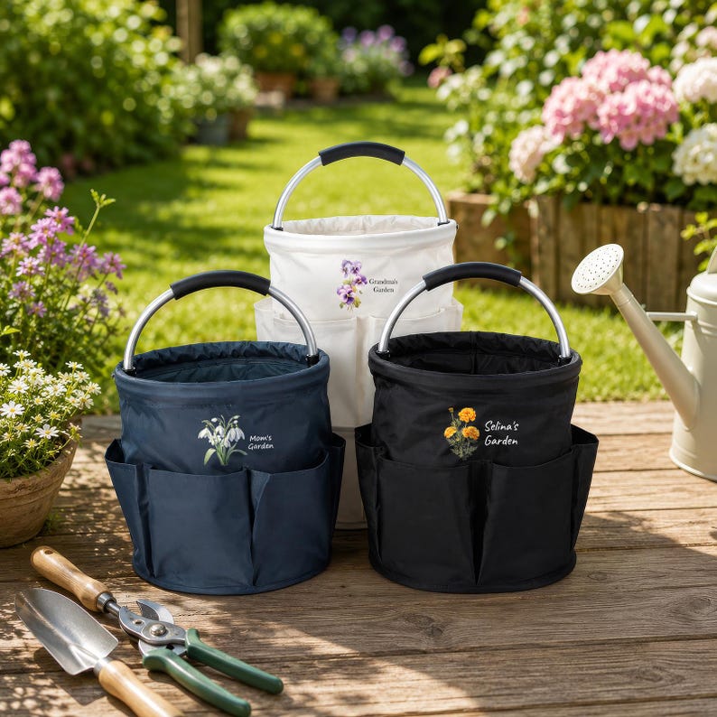 Puede incluir: Tres bolsas de jard&iacute;n cil&iacute;ndricas con asas de metal plateado y bolsillos exteriores. La bolsa azul marino muestra flores blancas y el texto "Mom's Garden", la blanca flores moradas y "Grandma's Garden", y la negra flores naranjas y "Selina's Garden".