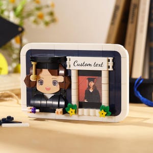 May include: A graduation-themed building block set featuring a cartoon graduate and a framed photo. The set includes a navy blue and white frame with the text "Custom text". The graduate figure is wearing a black cap and gown.