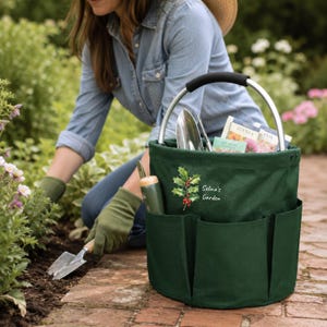 Puede incluir: Una bolsa cil&iacute;ndrica de color verde oscuro para herramientas de jard&iacute;n con asa de metal plateado y agarre negro. La bolsa cuenta con m&uacute;ltiples bolsillos exteriores, un gr&aacute;fico de acebo y el texto blanco "Selina's Garden".