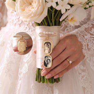 May include: A gold-toned metal locket pin attached to a bouquet handle. The locket features a hinged design displaying two black and white photographs of couples. The pin is adorned with four small white pearls and is secured to a cream satin ribbon.