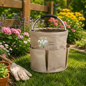 Puede incluir: Una bolsa de lona beige para herramientas de jard&iacute;n con asa de metal y varios bolsillos exteriores. El frente presenta una ilustraci&oacute;n de flores blancas y el texto negro Mom's Garden. La bolsa tiene forma cil&iacute;ndrica.