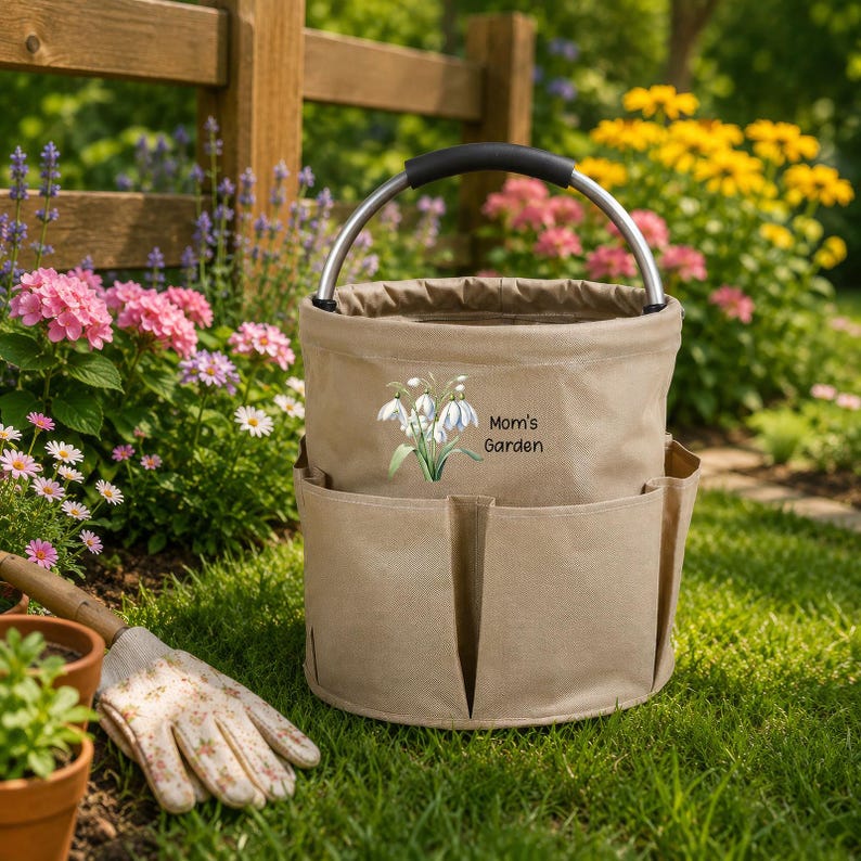 Puede incluir: Una bolsa de lona beige para herramientas de jard&iacute;n con asa de metal y varios bolsillos exteriores. El frente presenta una ilustraci&oacute;n de flores blancas y el texto negro Mom's Garden. La bolsa tiene forma cil&iacute;ndrica.