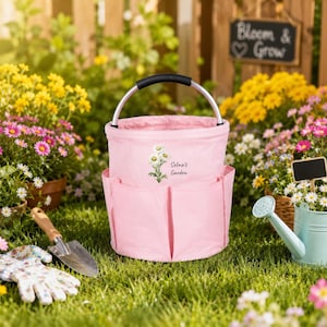 Puede incluir: Una bolsa de jard&iacute;n de tela rosa claro con un asa plateada y negra. La bolsa cuenta con m&uacute;ltiples bolsillos exteriores y un gr&aacute;fico impreso de flores de margarita blancas con el texto Selina's Garden en el panel frontal.
