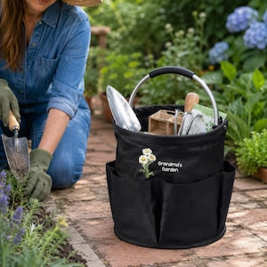 Puede incluir: Una bolsa de jard&iacute;n de tela negra con un asa de metal plateado y varios bolsillos exteriores. El frente presenta un gr&aacute;fico de margaritas bordado y el texto blanco Grandma's Garden. Contiene una pala y macetas.