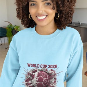 May include: A light blue sweatshirt featuring a soccer ball design with the text "WORLD CUP 2026 QATAR". The graphic is in maroon, white, and gray. The sweatshirt is made of a soft material.