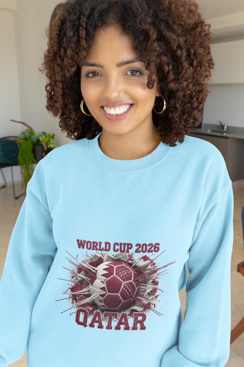 May include: A light blue sweatshirt featuring a soccer ball design with the text "WORLD CUP 2026 QATAR". The graphic is in maroon, white, and gray. The sweatshirt is made of a soft material.