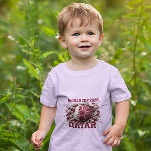 May include: A young child wearing a light purple t-shirt with the text "WORLD CUP 2026" and "QATAR" printed on the front, along with a soccer ball graphic. The child is standing in front of green foliage.