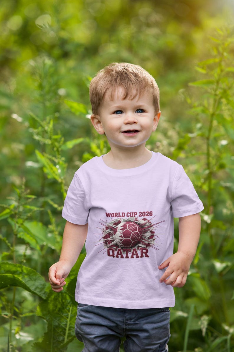 May include: A young child wearing a light purple t-shirt with the text "WORLD CUP 2026" and "QATAR" printed on the front, along with a soccer ball graphic. The child is standing in front of green foliage.