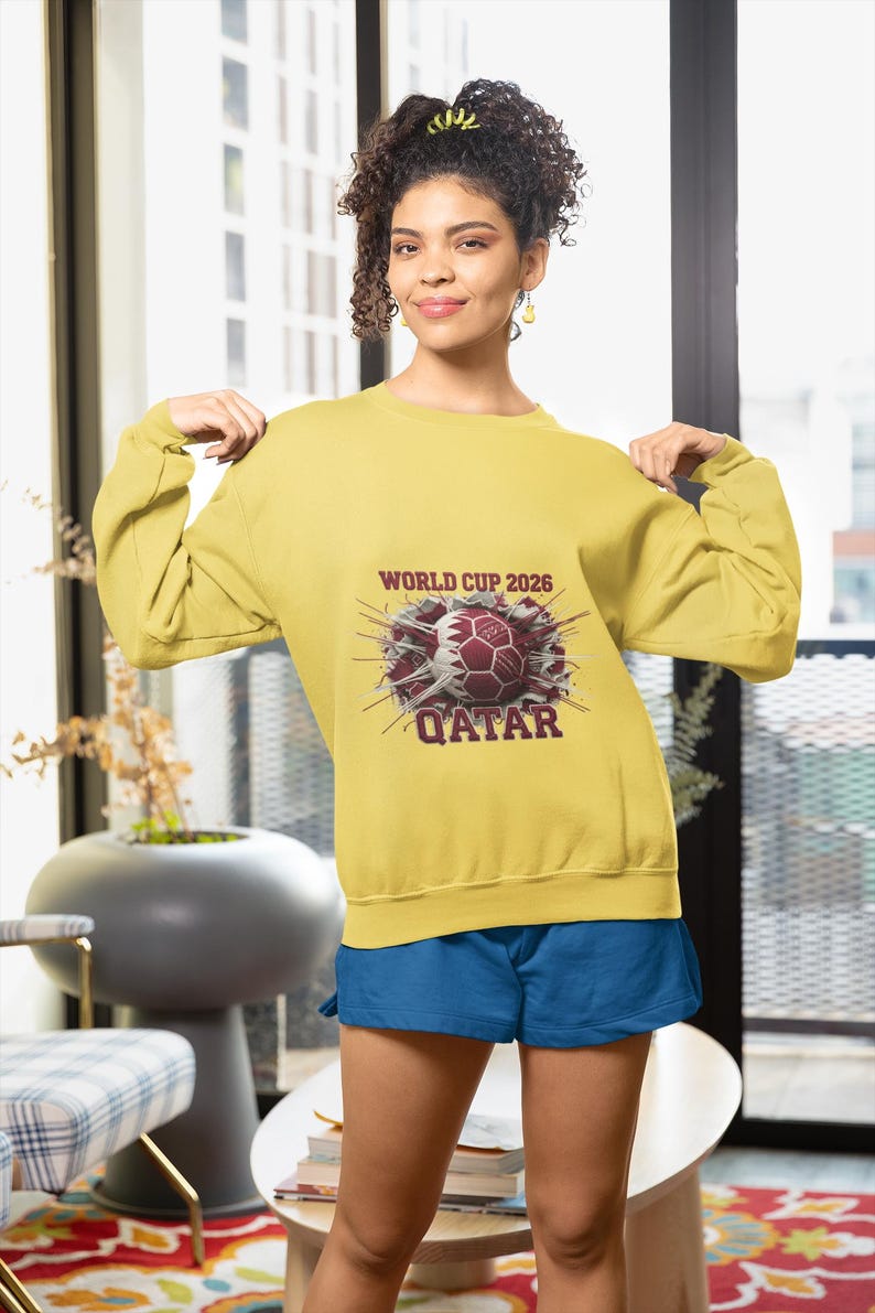 May include: Mustard yellow sweatshirt with a graphic of a soccer ball and the text "WORLD CUP 2026 QATAR". The sweatshirt is paired with blue shorts. The image is taken indoors.