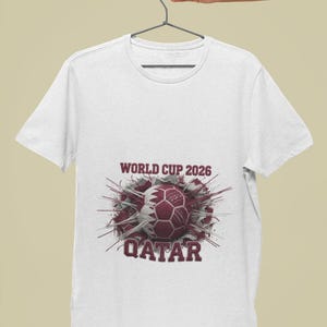 May include: White t-shirt featuring a maroon graphic of a soccer ball with the Qatar flag design. The text "WORLD CUP 2026" and "QATAR" are printed above and below the ball. The shirt is hanging on a gray hanger.