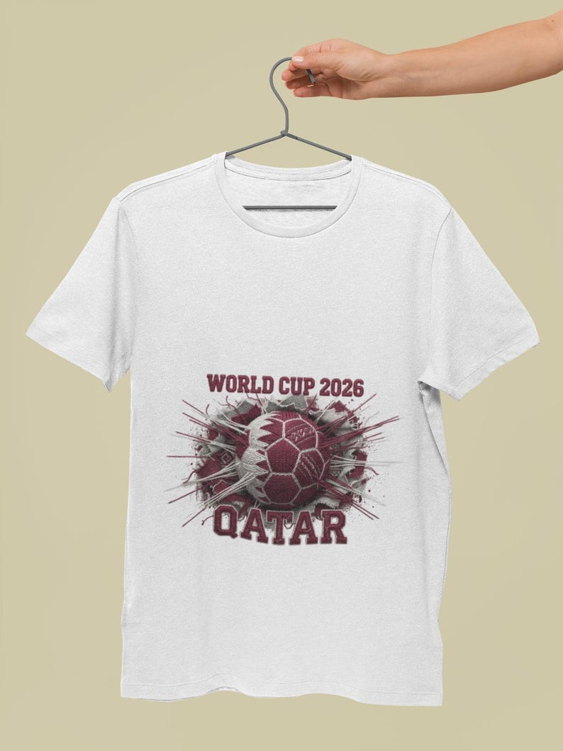 May include: White t-shirt featuring a maroon graphic of a soccer ball with the Qatar flag design. The text "WORLD CUP 2026" and "QATAR" are printed above and below the ball. The shirt is hanging on a gray hanger.