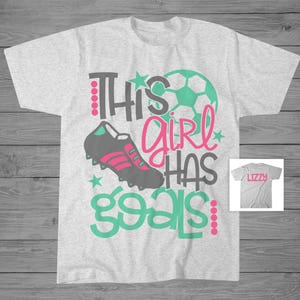 May include: Gray t-shirt with a green and pink design that says "This girl has goals". The design includes a soccer ball, a soccer cleat, and stars. The shirt is personalized with the name "Lizzy" on a separate gray t-shirt.