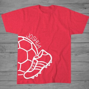 May include: Red t-shirt with a white soccer ball and a white shoe graphic. The text "JOSHUA" is written on the shirt.