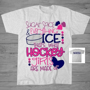 May include: A grey t-shirt with a pink and blue design that says "Sugar, Spice & Everything Nice: That's What Hockey Girls Are Made Of". The design also includes a hockey puck and a heart. The shirt is shown against a wooden background.